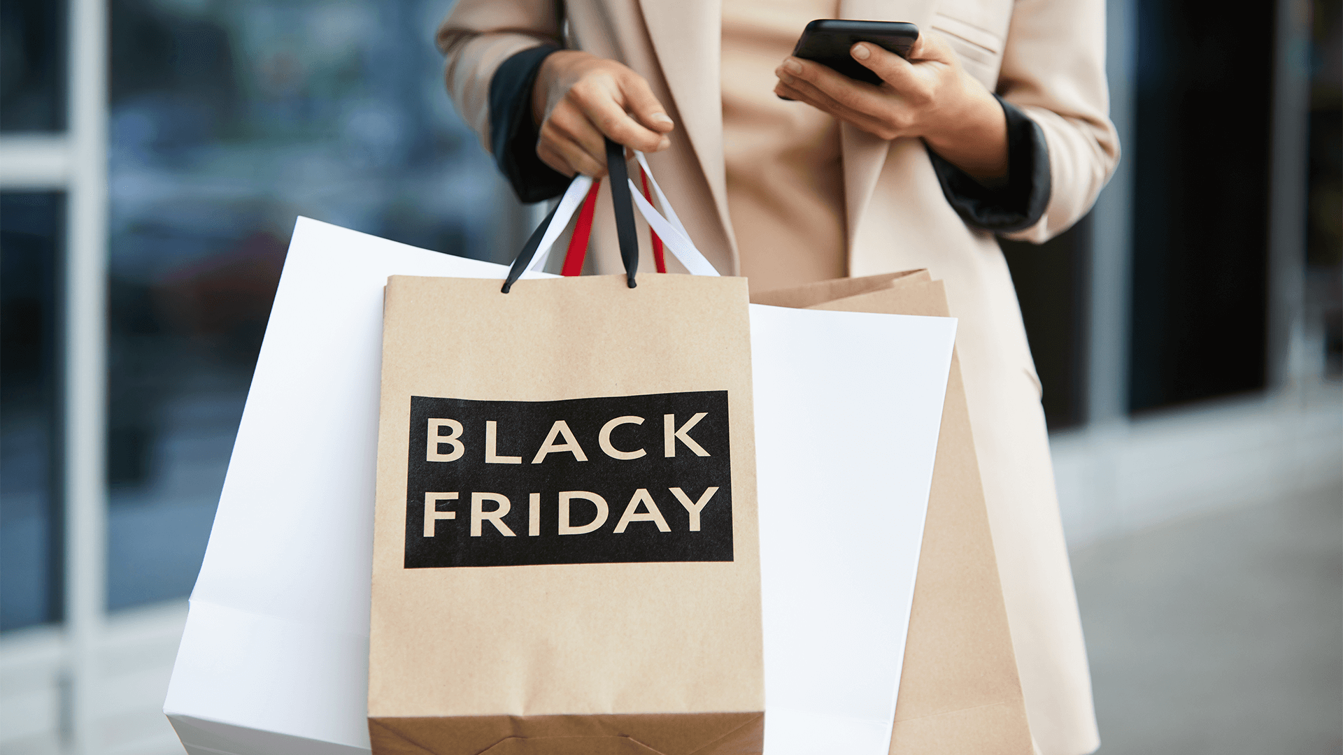 Mid-section portrait of stylish young woman holding shopping bags with Black Friday and texting on the go while leaving mall