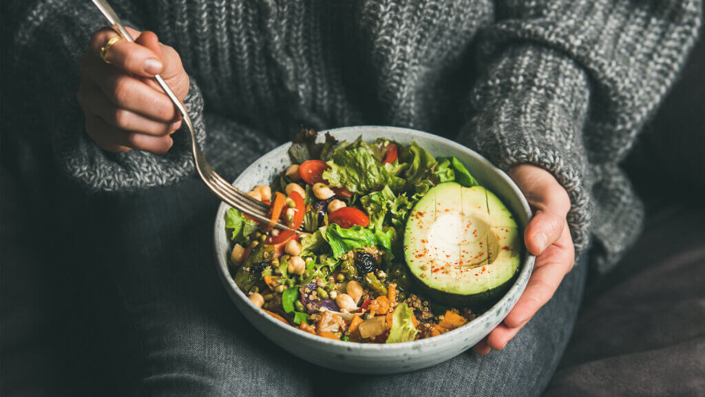 Woman in jeans and warm sweater holding bowl with fresh salad, avocado, grains, beans, roasted vegetables, close-up
